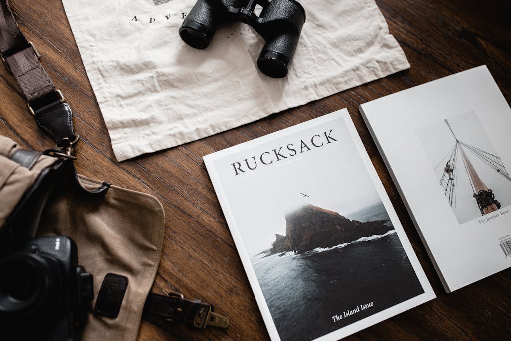 A stylish flat lay of travel essentials including binoculars and Rucksack magazine on a wooden surface.