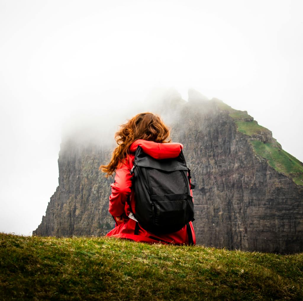 A woman sits with a backpack in front of fog-covered cliffs in the Faroe Islands, perfect for adventure travel themes.