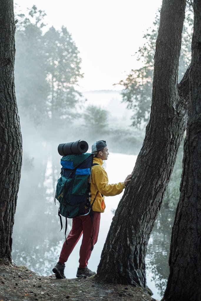 An adventurous hiker with backpack and gear standing in a misty forest by a tranquil river.